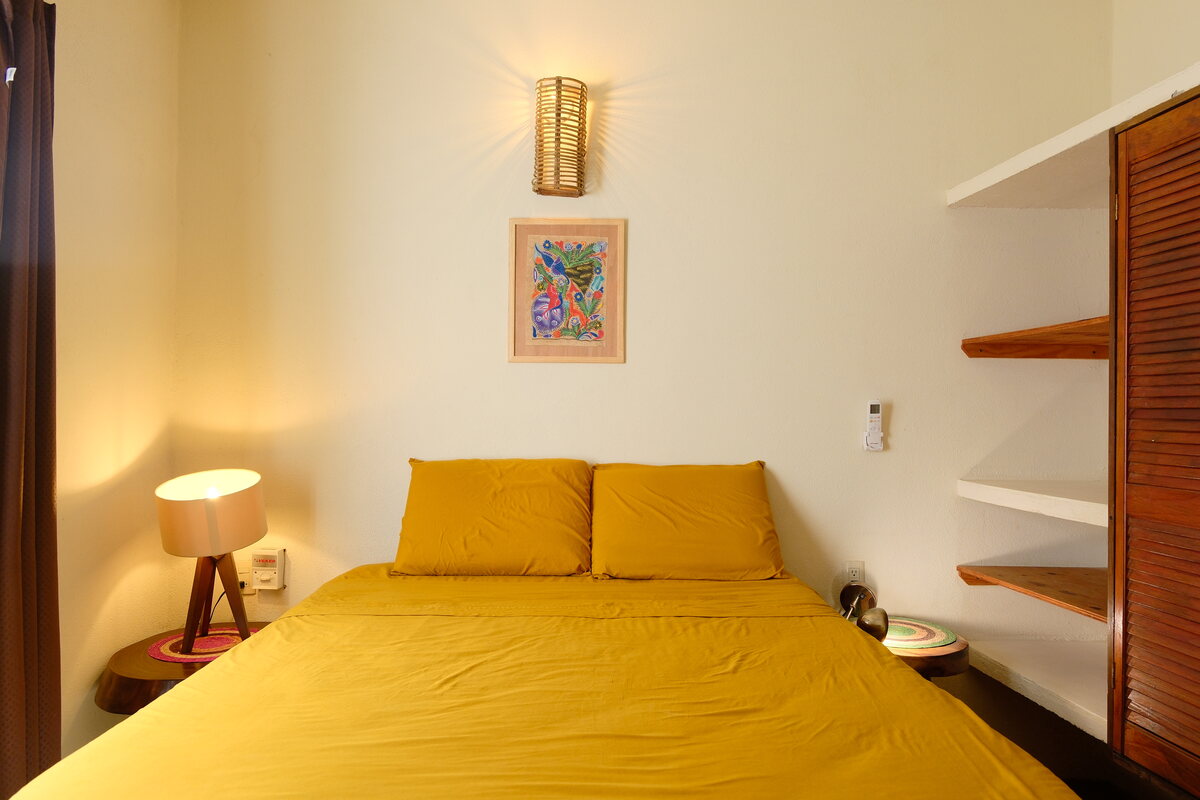 Casa Bel bedroom — made bed beneath a folk-art print, with a woven wall sconce and bedside lamp