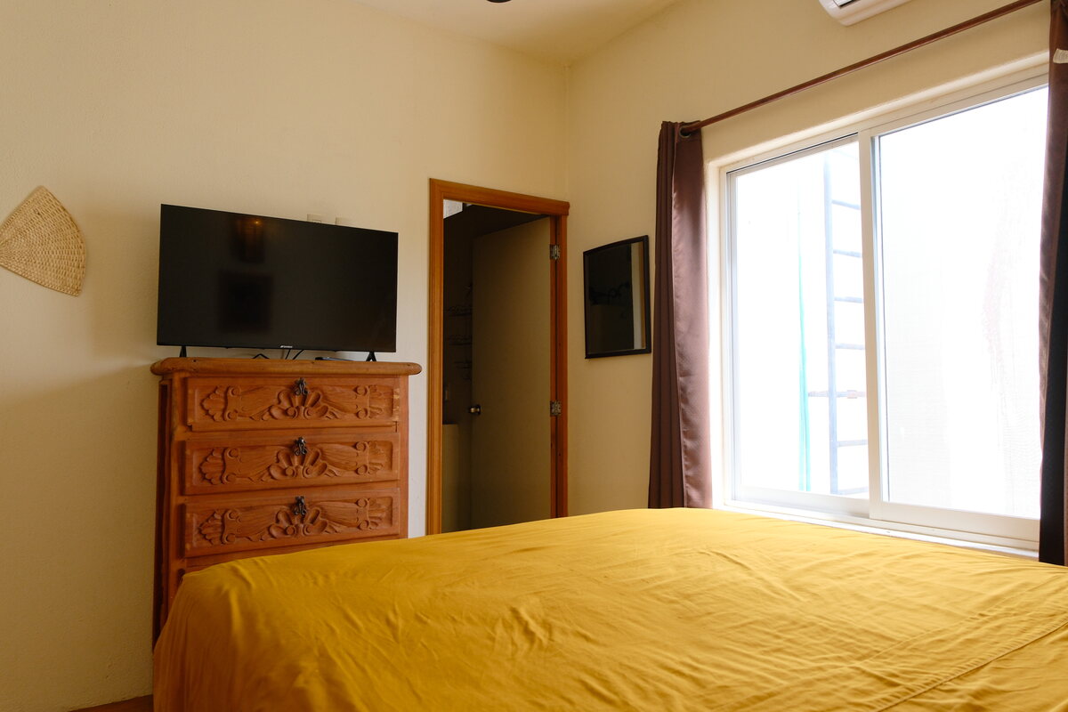 Casa Bel bedroom — flat-screen TV on a carved wood dresser, the door open to the en-suite bathroom, beside a sunlit window